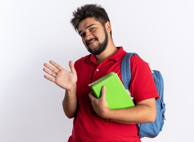 Young bearded student guy in red polo shirt with backpack holding notebooks  happy and cheerful smiling waving with hand standing over white wall