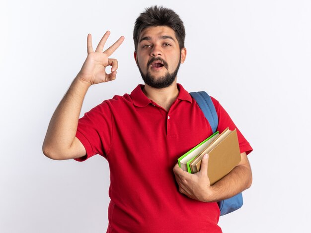 Молодой бородатый студент-парень в красной рубашке поло с рюкзаком, держащий ноутбуки, выглядит счастливым и позитивным, показывая знак ОК, стоящий над белой стеной