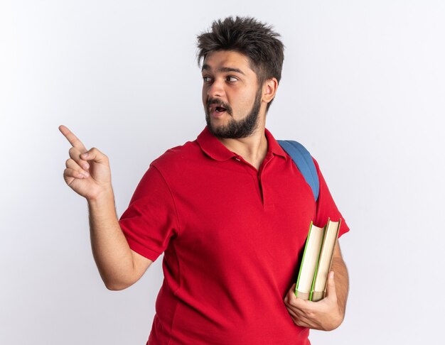 Young bearded student guy in red polo shirt with backpack holding notebooks looking aside pointing with index finger to the side smiling standing