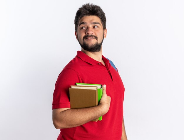 Young bearded student guy in red polo shirt with backpack holding notebooks looking aside smiling confident standing over white wall