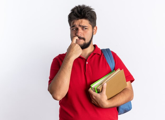 Free photo young bearded student guy in red polo shirt with backpack holding notebooks looking aside stressed and nervous biting nails standing over white wall