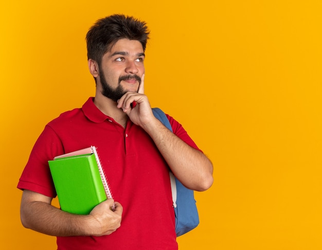 Молодой бородатый студент-парень в красной рубашке поло с рюкзаком держит ноутбуки, глядя в сторону с задумчивым выражением лица, думая позитивно, улыбаясь, стоя на оранжевом фоне