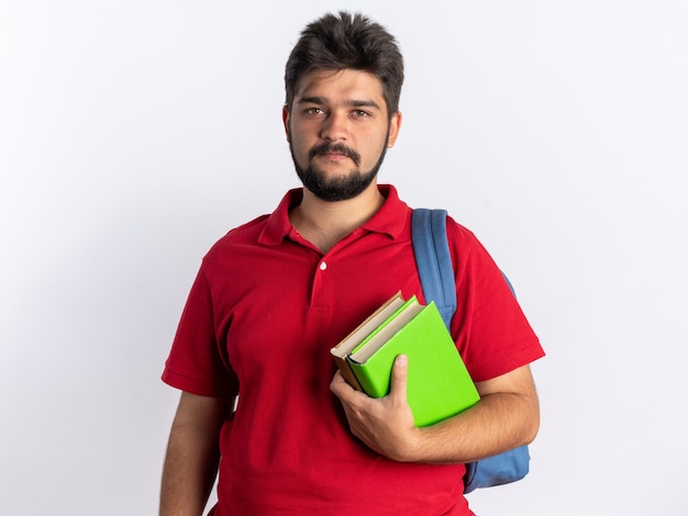 白い背景の上に立っている真剣な自信を持ってカメラを見ているノートブックを保持しているバックパックと赤いポロシャツの若いひげを生やした学生の男