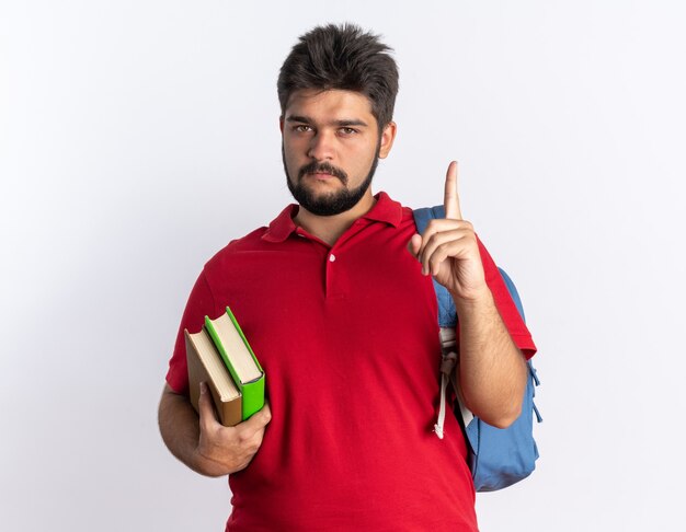 白い背景の上に立っている人差し指を示す深刻な顔でカメラを見てノートを保持しているバックパックと赤いポロシャツの若いひげを生やした学生の男