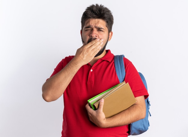 Молодой бородатый студент-парень в красной рубашке поло с рюкзаком, держащий ноутбуки, выглядит усталым и скучающим, зевая, стоя на белом фоне