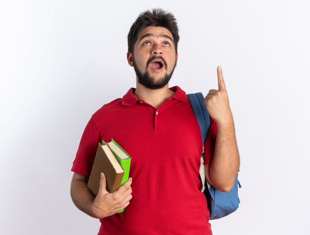 Young bearded student guy in red polo shirt with backpack holding notebooks looking up happy and excited pointing with index finger up standing over white wall