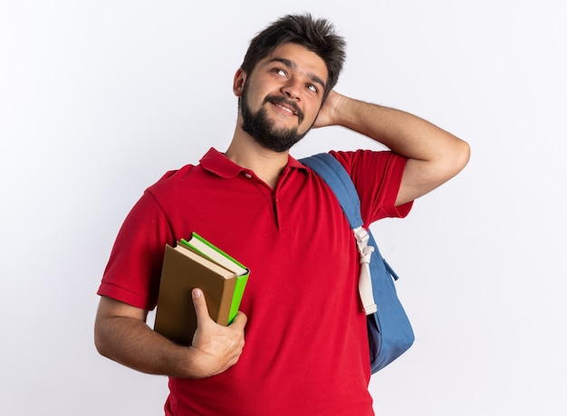 Young bearded student guy in red polo shirt with backpack holding notebooks looking up smiling thinking positive standing over white wall