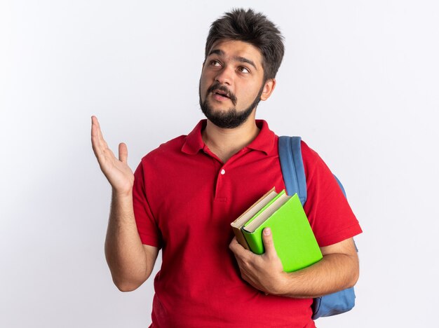 Young bearded student guy in red polo shirt with backpack holding notebooks looking up with smile on face with arm raised standing