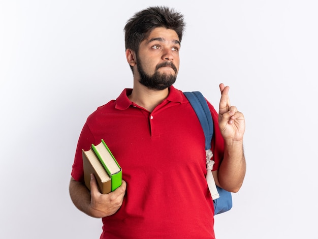 Молодой бородатый студент-парень в красной рубашке поло с рюкзаком держит тетради, загадывая желание с выражением надежды, скрещивая пальцы стоя