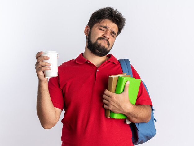 Young bearded student guy in red polo shirt with backpack holding notebooks and peper cup looking displeased and confused standing over white background