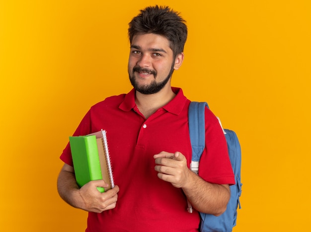 Молодой бородатый студент-парень в красной рубашке поло с рюкзаком держит тетради, указывая указательным пальцем на камеру, весело улыбаясь, стоя на оранжевом фоне