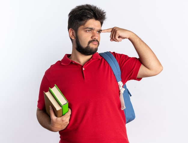 Молодой бородатый студент-парень в красной рубашке поло с рюкзаком держит тетради, указывая указательным пальцем на его висок, выглядит уверенно и думает стоя