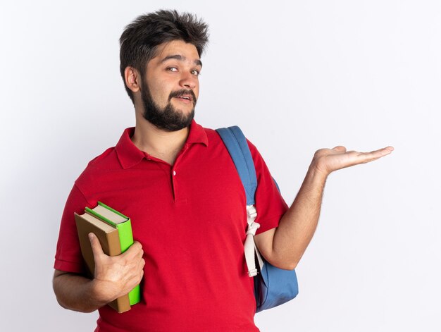 Young bearded student guy in red polo shirt with backpack holding notebooks presenting copy space with arm smiling cheerfully standing over white wall