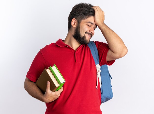 赤いポロシャツを着た若いひげを生やした学生の男が、ノートを持ったバックパックを持って、立っている彼の頭の上に手を置いて混乱している
