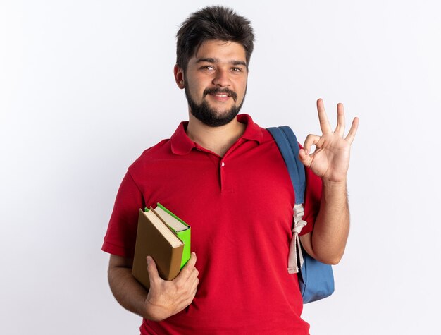 Young bearded student guy in red polo shirt with backpack holding notebooks  smiling showing ok sign standing over white wall