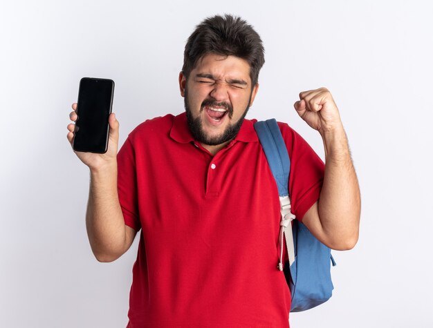 Молодой бородатый студент-парень в красной рубашке поло с рюкзаком держит смартфон, сжимая кулак, счастливый и взволнованный стоя