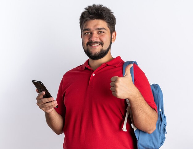 Молодой бородатый студент-парень в красной рубашке поло с рюкзаком держит смартфон, весело улыбаясь, показывая большие пальцы вверх стоя
