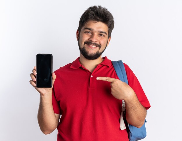 Young bearded student guy in red polo shirt with backpack holding smartphone pointing with idex finger at it smiling cheerfully standing