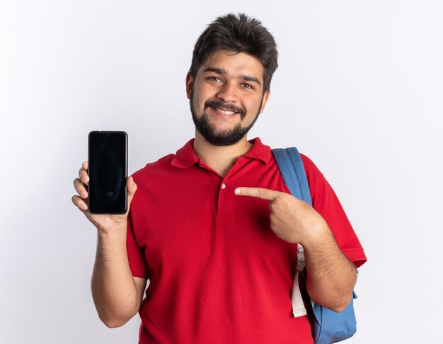 赤いポロシャツを着た若いひげを生やした学生の男が、スマートフォンを持ったバックパックを持ち、アイデックスの指でそれを指し、陽気に笑って立っている