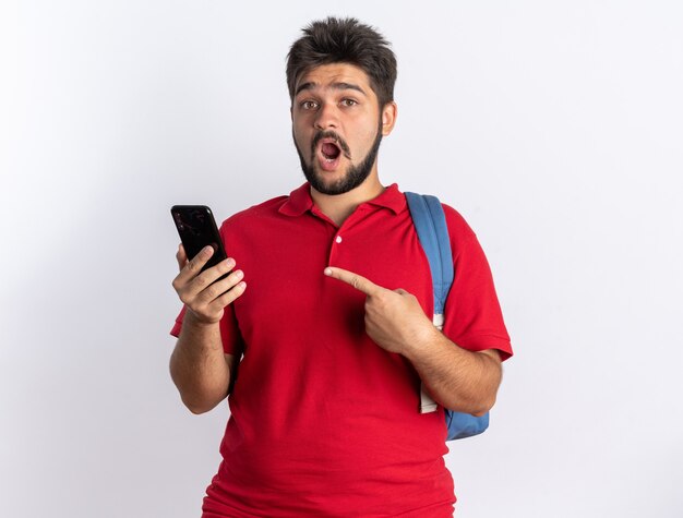 Young bearded student guy in red polo shirt with backpack holding smartphone pointing with index finger at it looking surprised standing