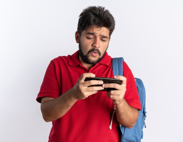 白い壁の上に立って心配しているスマートフォンを使ってゲームをするバックパックを持つ赤いポロシャツを着た若いひげを生やした学生の男