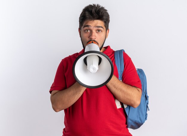 Молодой бородатый студент-парень в красной рубашке поло с рюкзаком кричит в мегафон, эмоциональный и взволнованный стоя