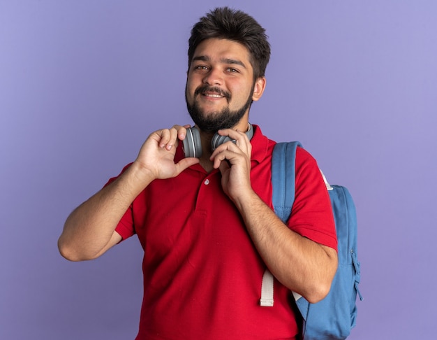 Молодой бородатый студент-парень в красной рубашке-поло с рюкзаком с наушниками, улыбаясь, весело и счастливо улыбается