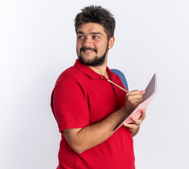 Young bearded student guy in red polo shirt with backpack writing in notebook looking confident smiling standing over white wall