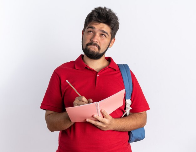 Young bearded student guy in red polo shirt with backpack writing in notebook looking up puzzled standing over white wall