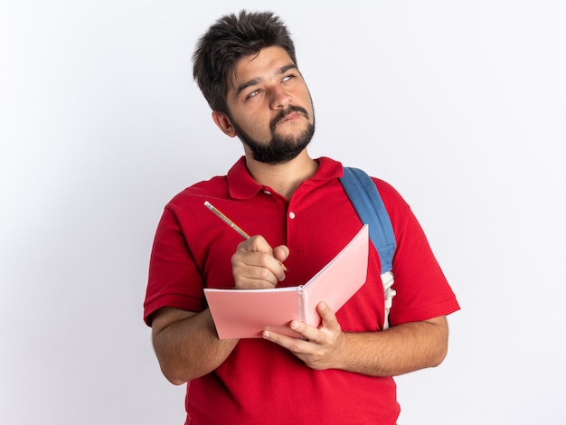 Young bearded student guy in red polo shirt with backpack writing in notebook looking up with pensive expression thinking standing