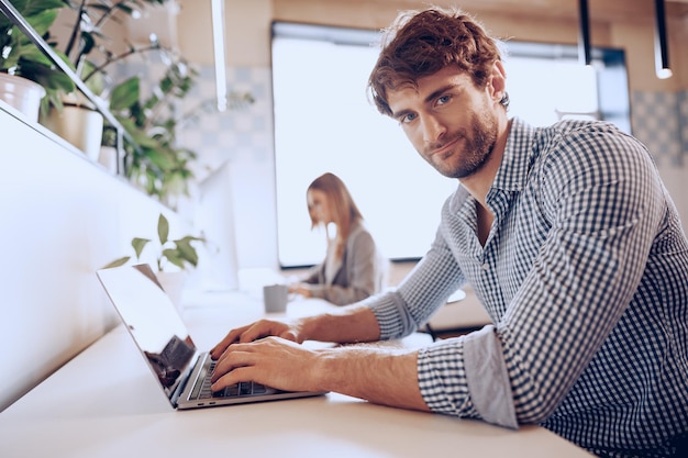 Молодой бородатый успешный бизнесмен, работающий на ноутбуке в офисе с коллегой-женщиной