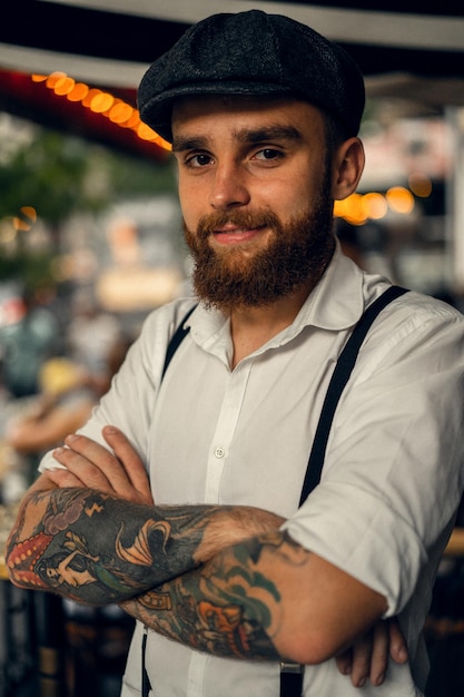 와인 한 잔과 함께 거리에 있는 카페에서 문신을 한 젊은 남자. 도시의 하얀 모자와 멜빵을 쓴 로맨틱한 남자. 피키 블라인더스. 오래 된 유행 복고풍입니다.