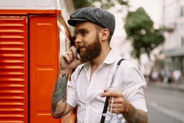 Young bearded tattooed man portrait close-up calls on the phone on the street. A romantic guy in a white shirt, cap and suspenders. Peaky Blinders. old-fashioned, retro.