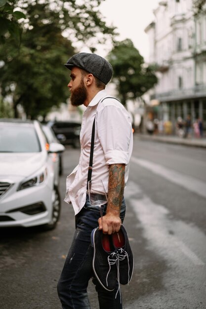 Young bearded tattooed man. A romantic guy in a white shirt, cap and suspenders walks in the city. revolves around a lamppost. Peaky Blinders. old-fashioned, retro.