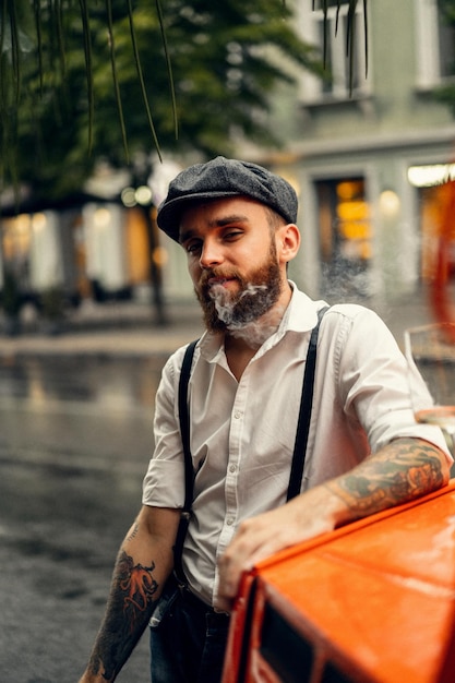 通りのカフェで若いひげを生やした入れ墨の男はタバコを吸います。街の白いシャツのキャップとサスペンダーのロマンチックな男。ピーキーブラインダーズ。古いファッショナブルなレトロ。