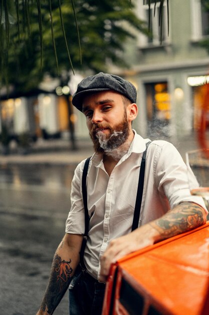 거리에 있는 카페에서 수염을 기른 젊은 문신남이 담배를 피우고 있다. 도시에서 흰색 셔츠 모자와 멜빵에 로맨틱 남자. 피키 블라인더스. 오래 된 유행 복고풍입니다.
