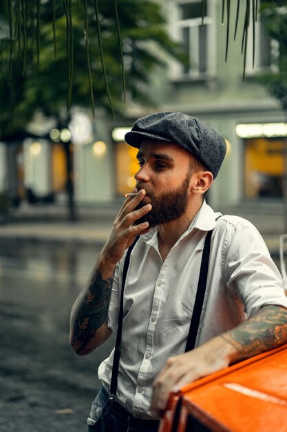 거리에 있는 카페에서 수염을 기른 젊은 문신남이 담배를 피우고 있다. 도시에서 흰색 셔츠 모자와 멜빵에 로맨틱 남자. 피키 블라인더스. 오래 된 유행 복고풍입니다.