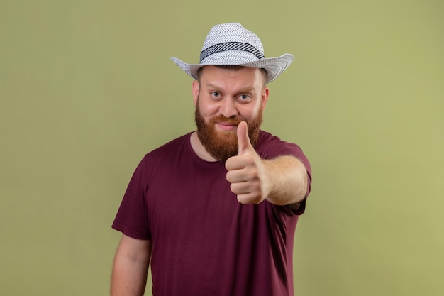 Young bearded traveler man in summer hat looking at camera smiling positive and happy showing thumbs up