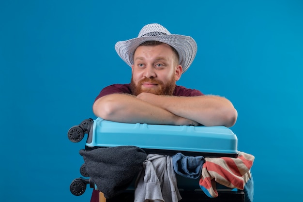 Young bearded traveler man in summer hat with suitcase full of clothes optimistic and happy smiling looking aside with dreamy look