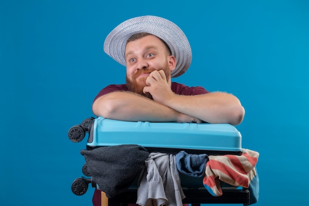 Молодой бородатый путешественник в летней шляпе с чемоданом, полным одежды, оптимистичный и счастливый улыбающийся, смотрящий в сторону с задумчивым выражением лица