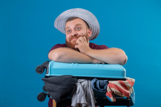 Young bearded traveler man in summer hat  with suitcase full of clothes optimistic and happy smiling looking aside with pensive expression on face 