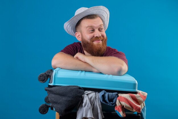 楽観的で幸せな笑顔の服でいっぱいのスーツケースと夏の帽子の若いひげを生やした旅行者の男