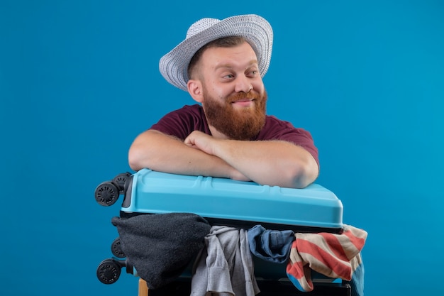 Young bearded traveler man in summer hat  with suitcase full of clothes optimistic and happy smiling looking aside 