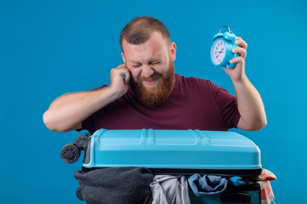 Young bearded traveler man  with suitcase full of clothes holding alarm clock covering his ear with finger with annoyed expression 