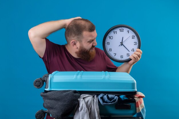 Young bearded traveler man  with suitcase full of clothes holding wall clock looking at it confused scratching his head 