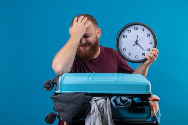 Young bearded traveler man  with suitcase full of clothes holding wall clock touching his head for mistake looking confused 