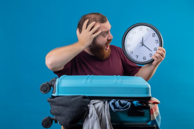 Uomo giovane viaggiatore barbuto con la valigia piena di vestiti che tengono l'orologio di parete che tocca la sua testa nel panico