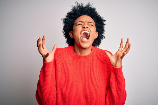 Young beautiful African American afro woman with curly hair wearing red casual sweater celebrating mad and crazy for success with arms raised and closed eyes screaming excited Winner concept