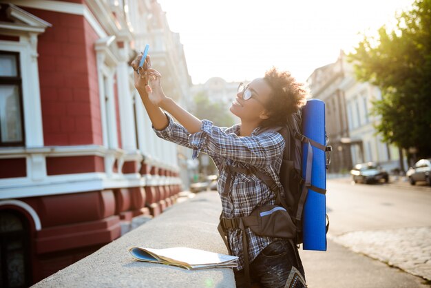 Young beautiful african female traveler with backpack smiling, making selfie.