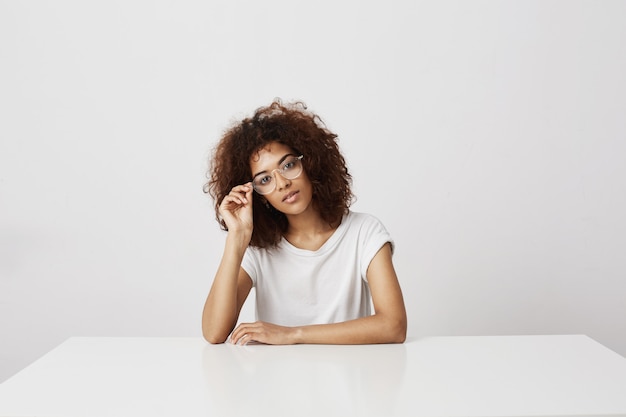 Young beautiful african girl in glasses smiling  sitting over white wall